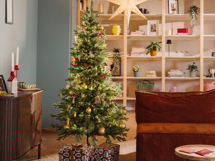 Die künstliche Nordmanntanne FairyTrees FT05 steht festlich geschmückt im Wohnzimmer einer kleinen Wohnung an einem winterlichen Abend.