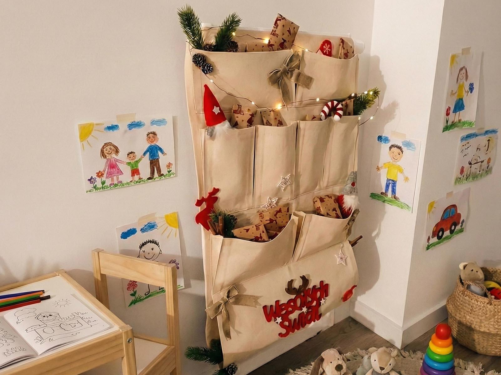 Der Adventskalender aus Stoff, geschm&uuml;ckt mit Zweigen des k&uuml;nstlichen Weihnachtsbaums FairyTrees und Lichtern, h&auml;ngt in der Ecke des Kinderzimmers.
