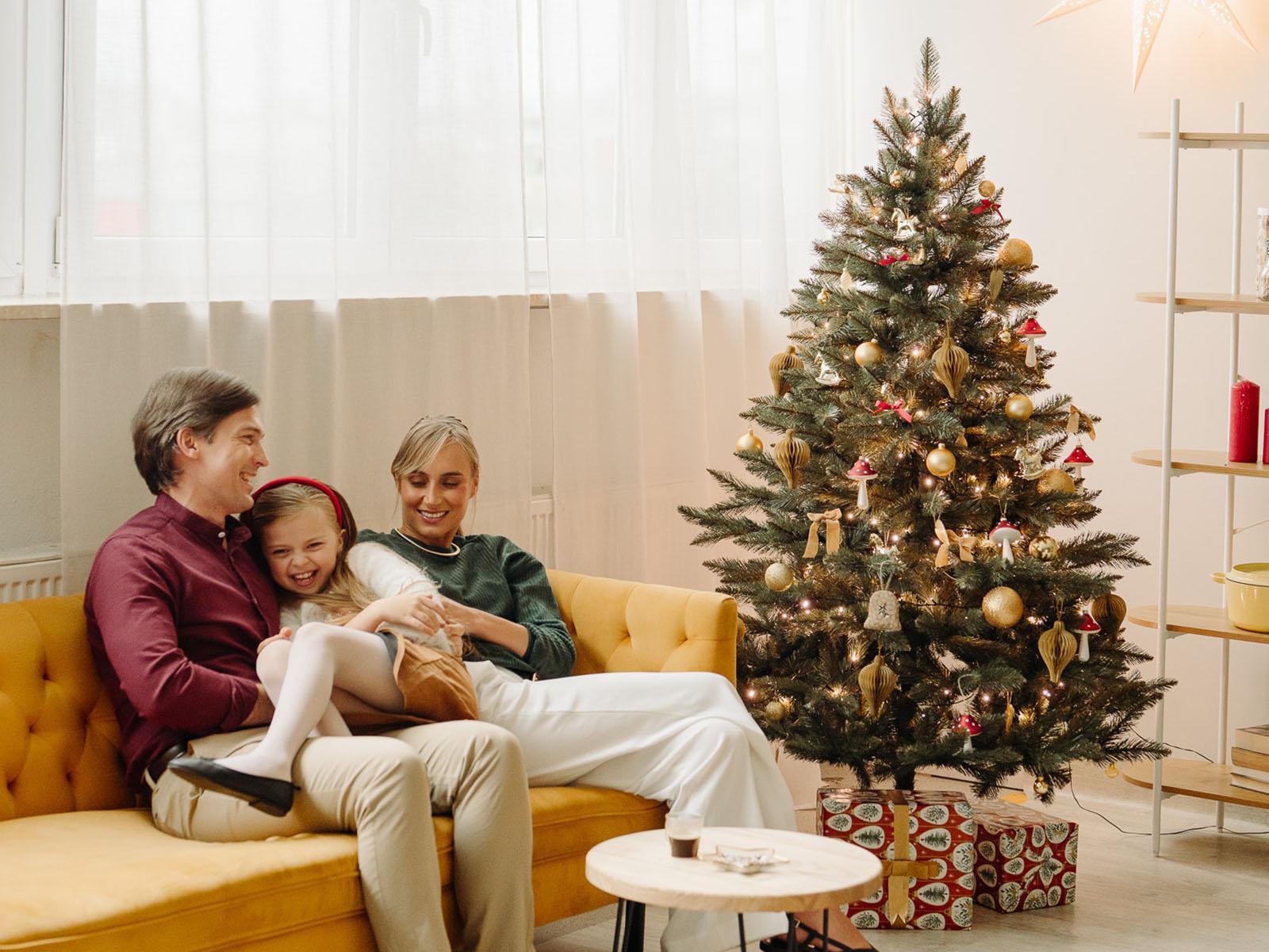 Mutter und Vater spielen mit ihrer Tochter auf dem Sofa neben der k&uuml;nstlichen Nordmanntanne FairyTrees FT14 mit dichten PVC-Nadeln in einem minimalistischen, farbenfrohen und modernen Wohnzimmer.