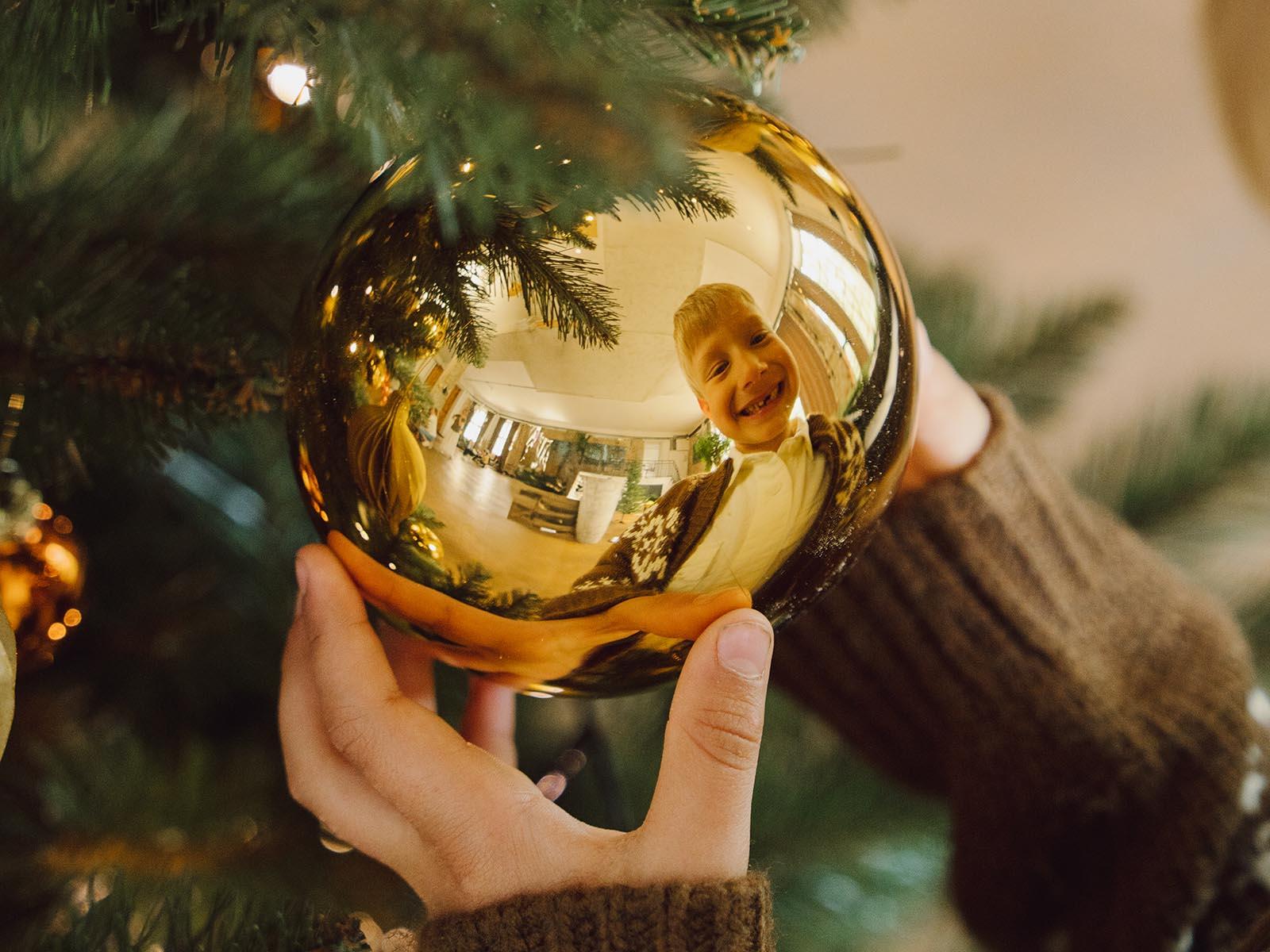 Ein kleiner Junge hält eine goldene Kugel an einem künstlichen Weihnachtsbaum FairyTrees FT14 fest, und in der Reflexion sieht man sein Lächeln.