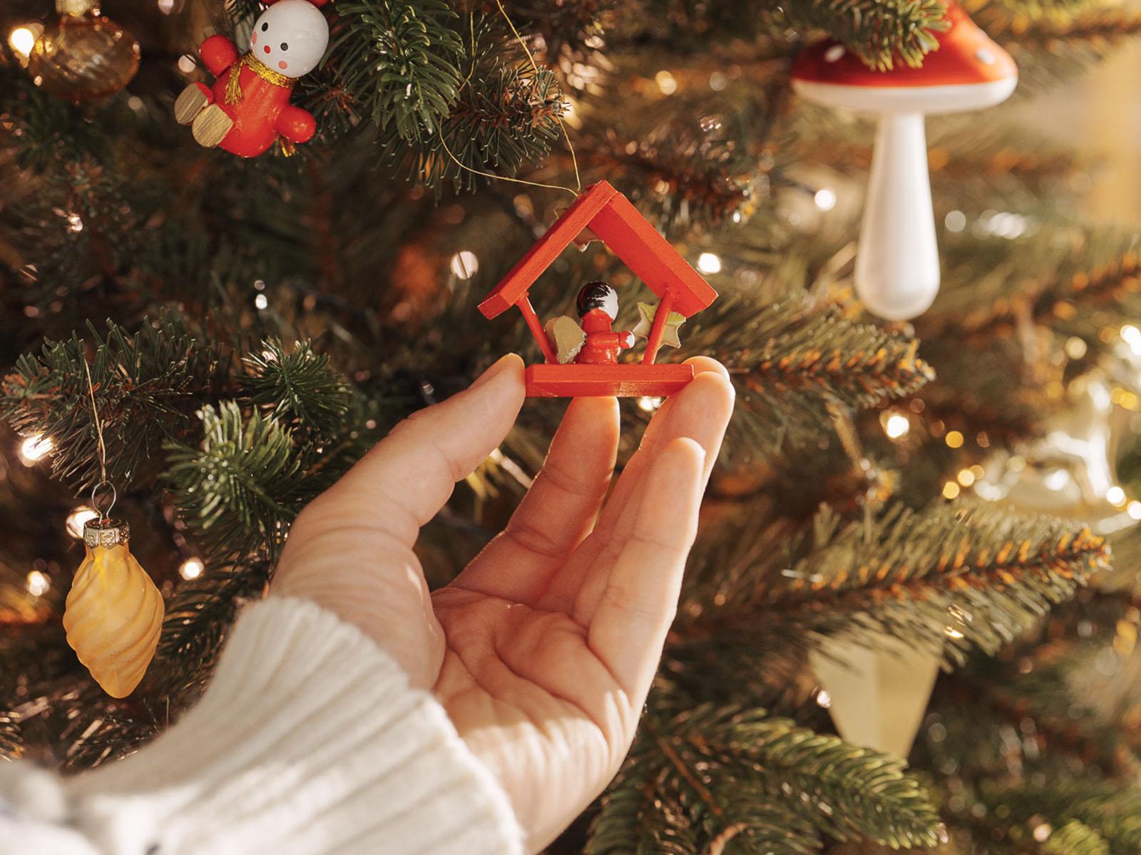 Die Hand einer Frau h&auml;ngt einen Holzanh&auml;nger an die festlich geschm&uuml;ckte k&uuml;nstliche Nordmanntanne FairyTrees FT29 mit PE- und PVC-Nadeln.