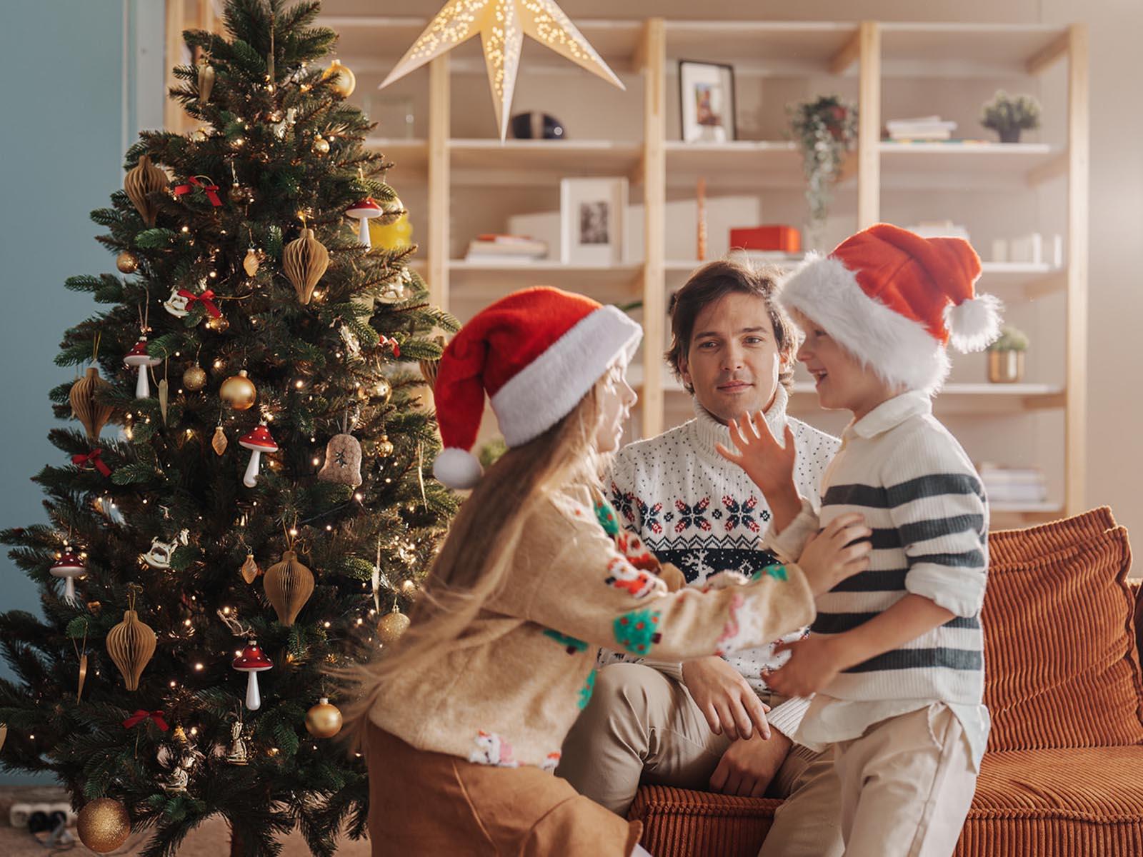 Der Vater sitzt erschöpft auf dem Sofa, während die Kinder mit Weihnachtsmützen am künstlichen Weihnachtsbaum FairyTrees FT29 herumtollen.
