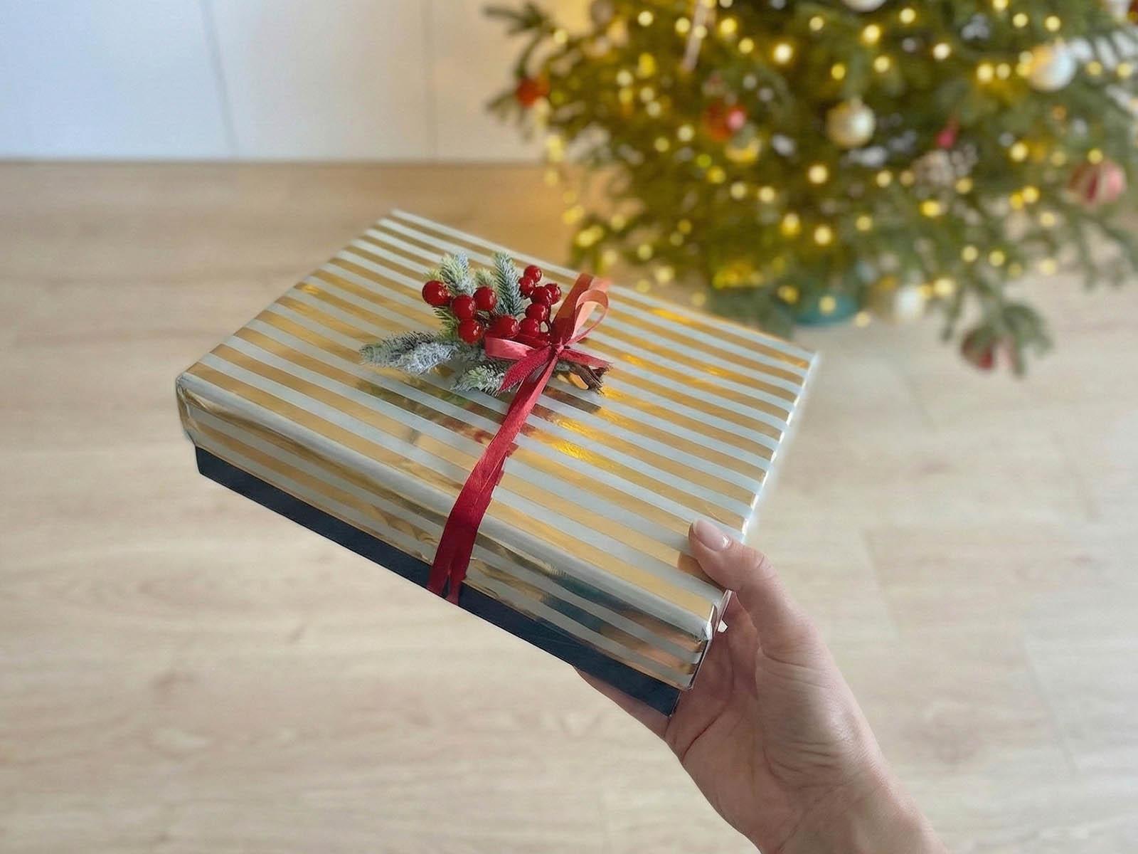 Eine Frau h&auml;lt ein verpacktes Geschenk in der Hand, das mit einem roten Band verziert und mit Zweigen eines k&uuml;nstlichen FairyTrees-Weihnachtsbaums dekoriert ist; im Hintergrund ist ein beleuchteter FairyTrees-Weihnachtsbaum zu sehen.