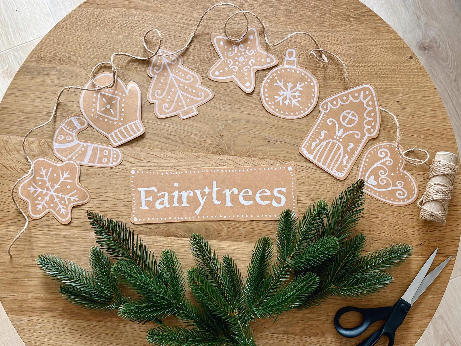 Die Zweige des künstlichen Weihnachtsbaums von FairyTrees liegen zusammen mit Papierdekorationen im Lebkuchenstil auf einer Holzplatte – bereit für die Herstellung von Girlanden.