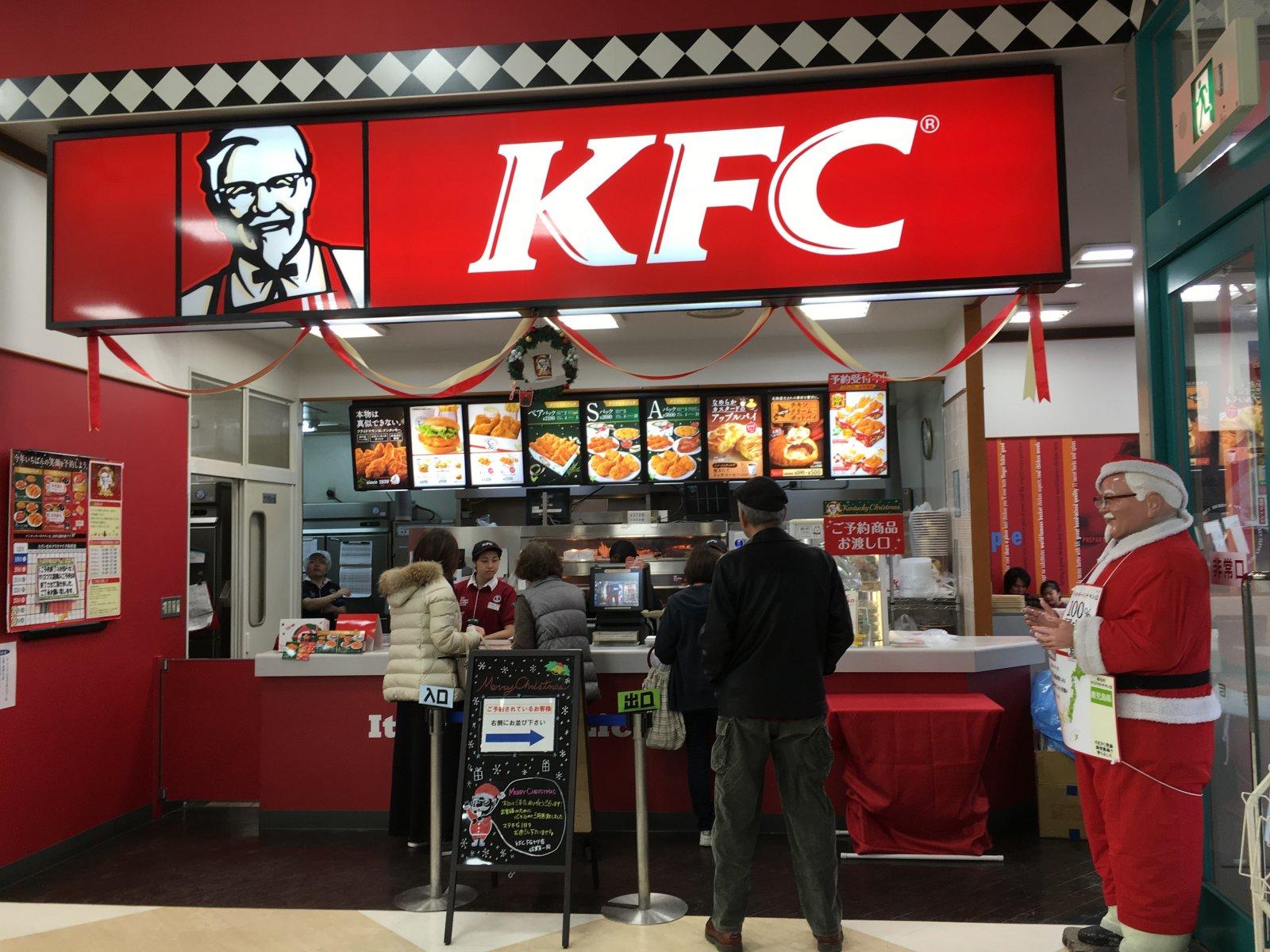 Kunden bestellen Essen an der Theke eines weihnachtlich dekorierten KFC-Restaurants, daneben steht eine als Weihnachtsmann verkleidete Person.