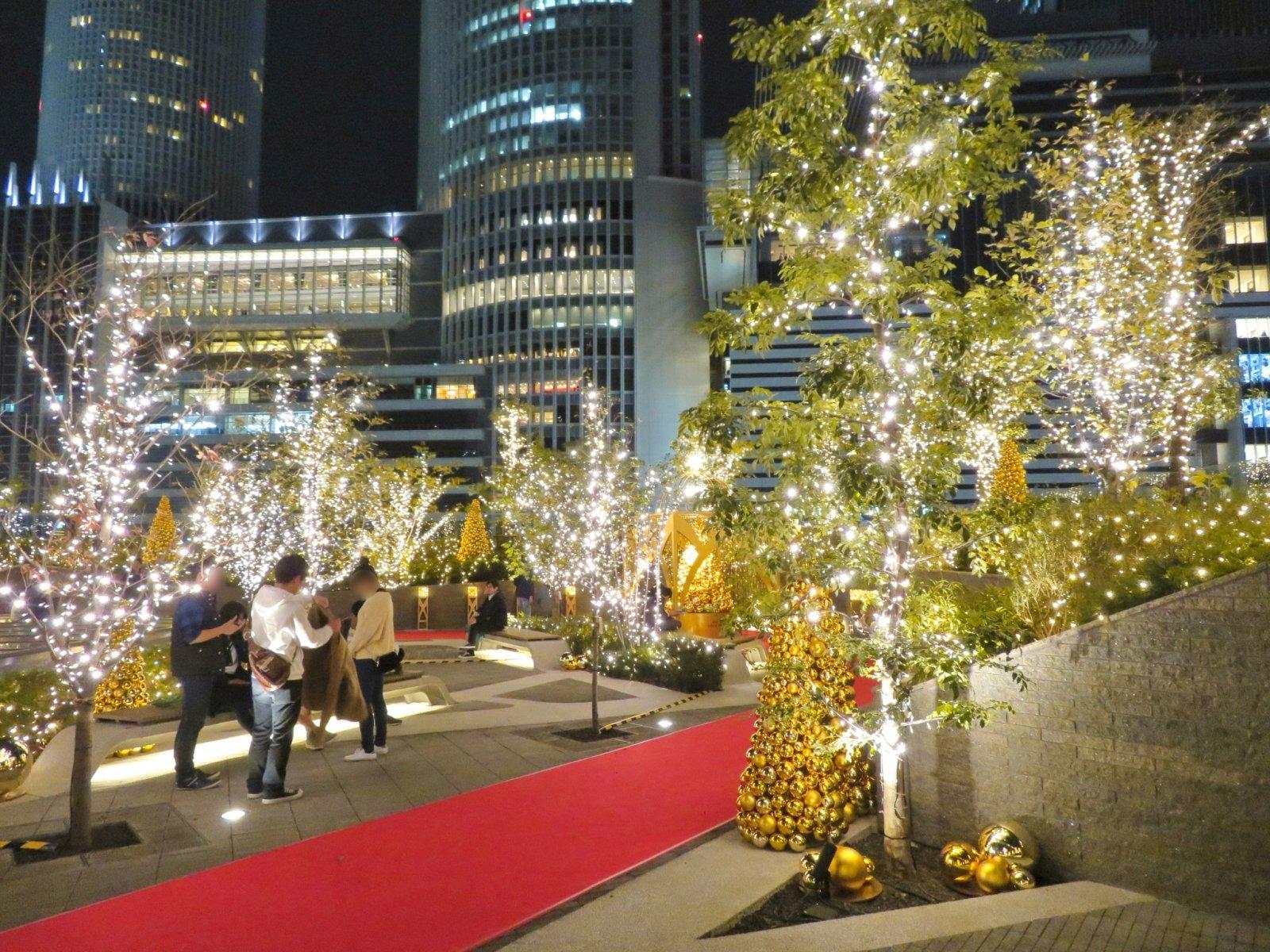 Eine Dachterrasse des Dai-Nagoya-Geb&auml;udes in Tokio ist mit festlichen Lichtern und Weihnachtsb&auml;umen dekoriert, umgeben von moderner Architektur.