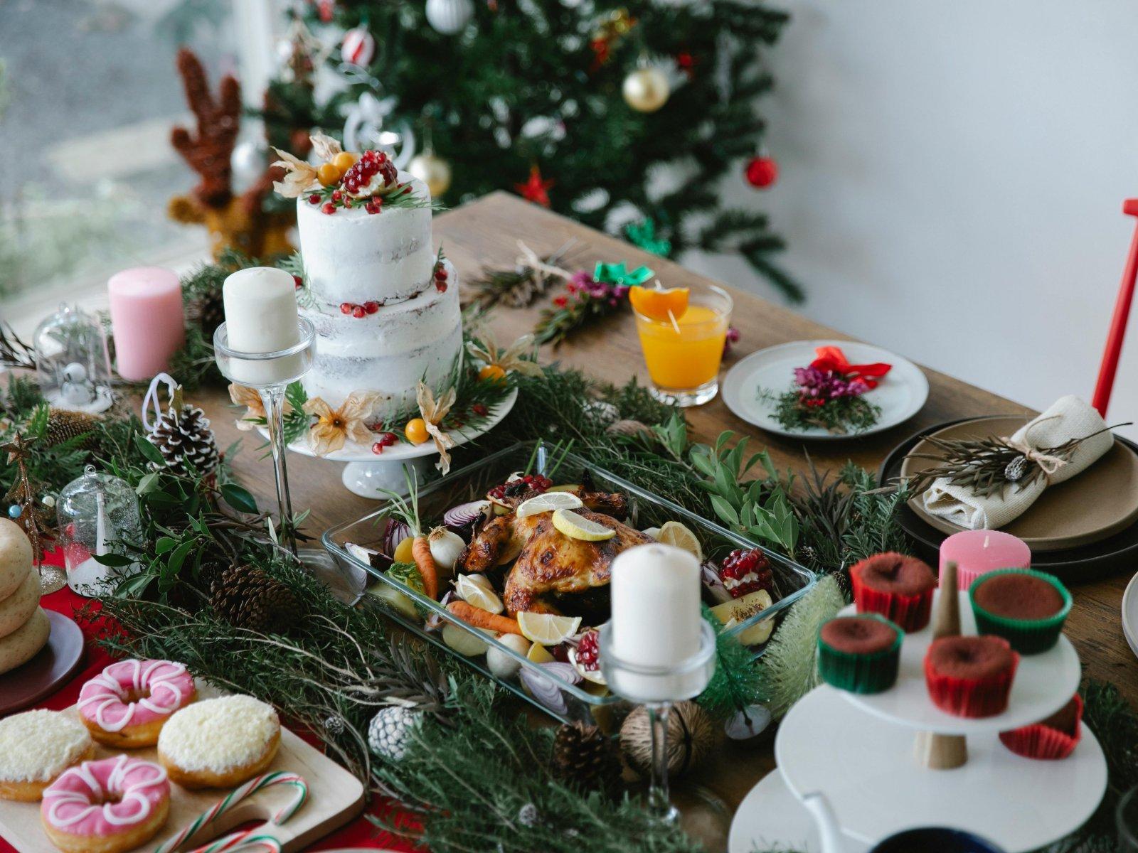 Auf dem Tisch stehen gebratenes Hähnchen, ein Weihnachtstorte und festliche Dekorationen, im Hintergrund ist ein künstlicher Weihnachtsbaum mit Kugeln zu sehen.